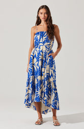 A woman is wearing a sleeveless, ankle-length dress with thin spaghetti straps. The dress features a bold, abstract tropical print in shades of blue and cream. It has a loose, flowing fit with a gathered bodice and a waistband that ties at the side. She is also wearing open-toe, strappy sandals in a light color.